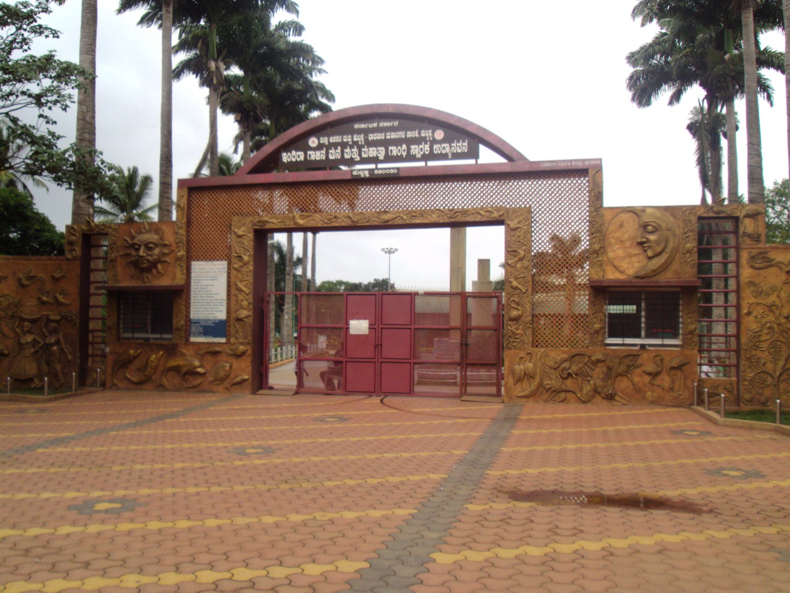 Ornate main entrance to Indira Gandhi Glass House Garden, Hubli, with sun and moon relief panels and palm trees behind.
